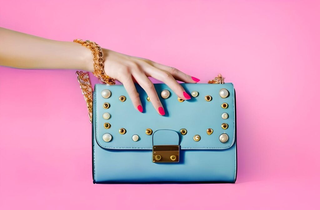 Hand with bright pink nails and gold bracelet holding a light blue studded handbag against a bold pink background